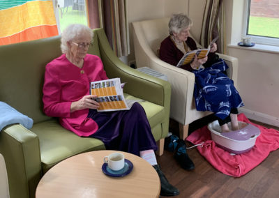 Two lady residents reading and relaxing in chairs together, with one having a foot spa