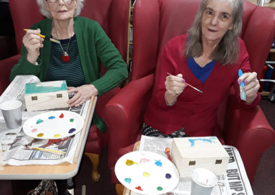 Residents sitting together painting wooden treasure boxes with colourful paints at Lulworth House Residential Care Home