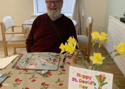 Male resident siting at a table with a vase of daffodils and his St Davids Day picture he'd coloured