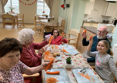 Residents and a young female relative sitting around a table peeling carrots to be used for lunch