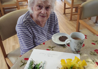 Female resident sitting at a table with a spring crafts picture and a cuppa and biscuit